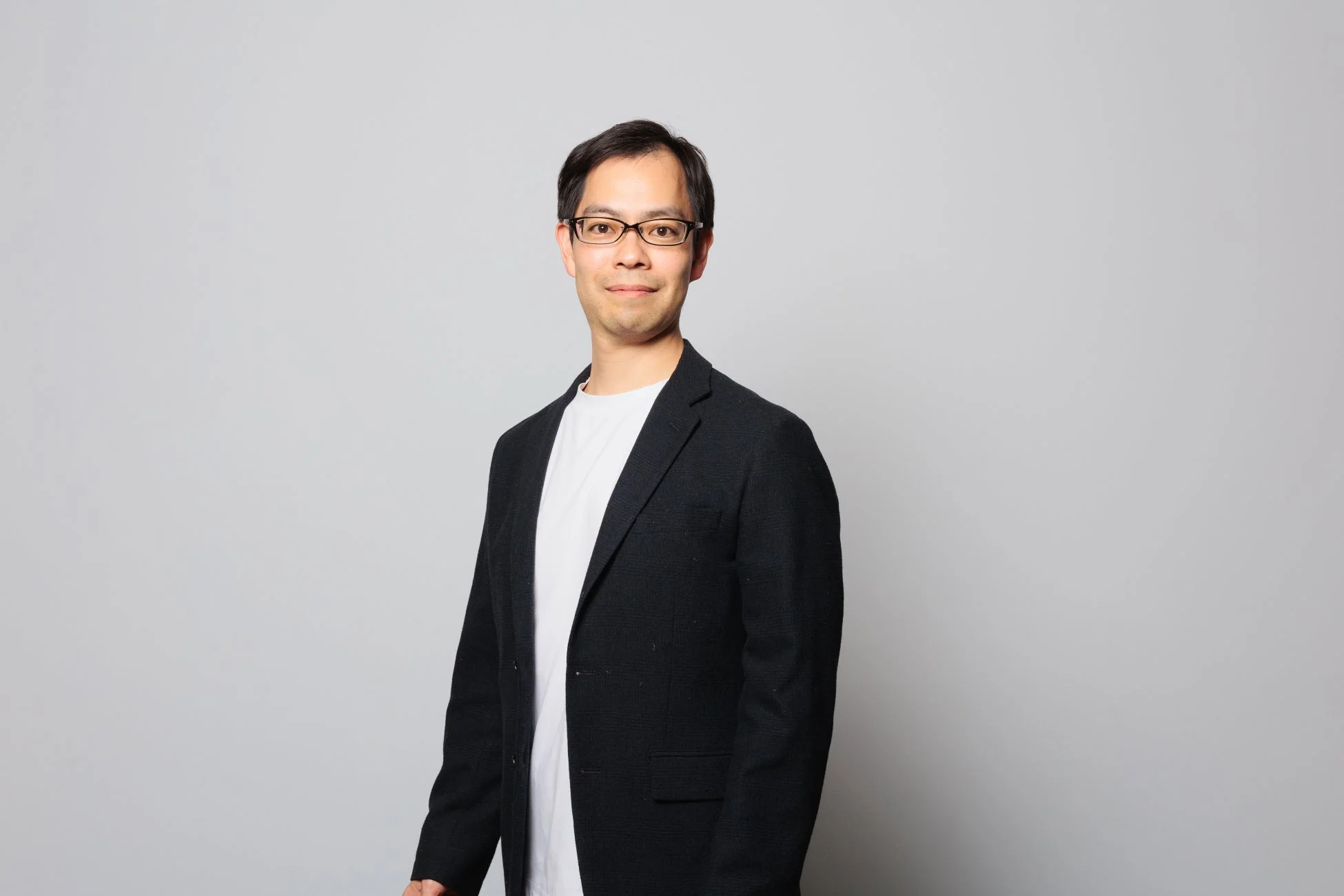 明るい背景を背に、眼鏡をかけたアジア系の男性が黒いジャケットと白いTシャツを着て立っています。カメラに向かって穏やかに微笑んでおり、上半身が写ったポートレート写真です。