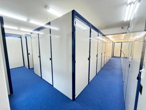 青いカーペットが敷かれた通路に、多数の白い扉のレンタル収納ユニットが整然と並ぶ屋内空間