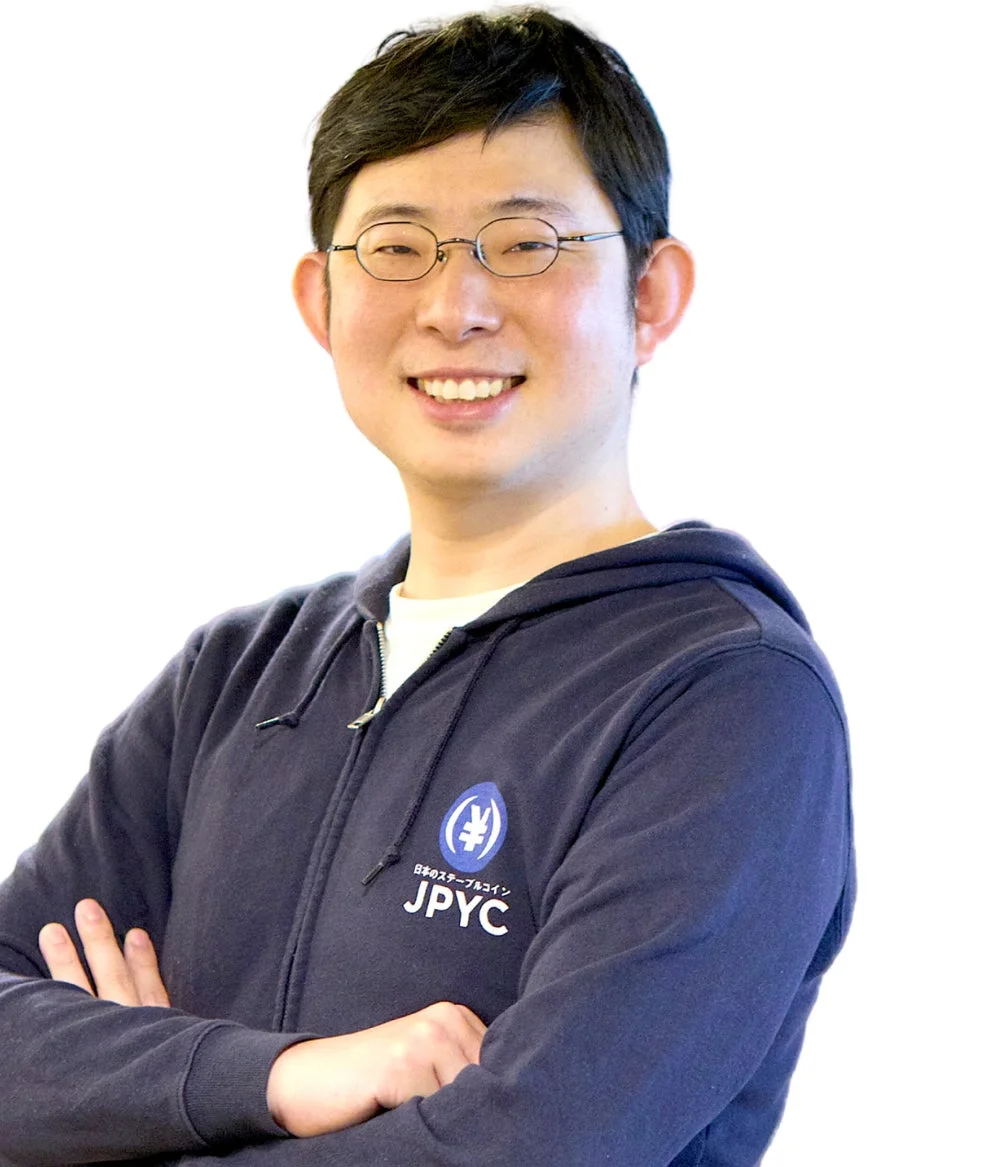 紺色のJPYCロゴ入りパーカーを着た眼鏡のアジア人男性が、腕組みをして笑顔で立っています。日本のステーブルコインに関連する人物のようです。