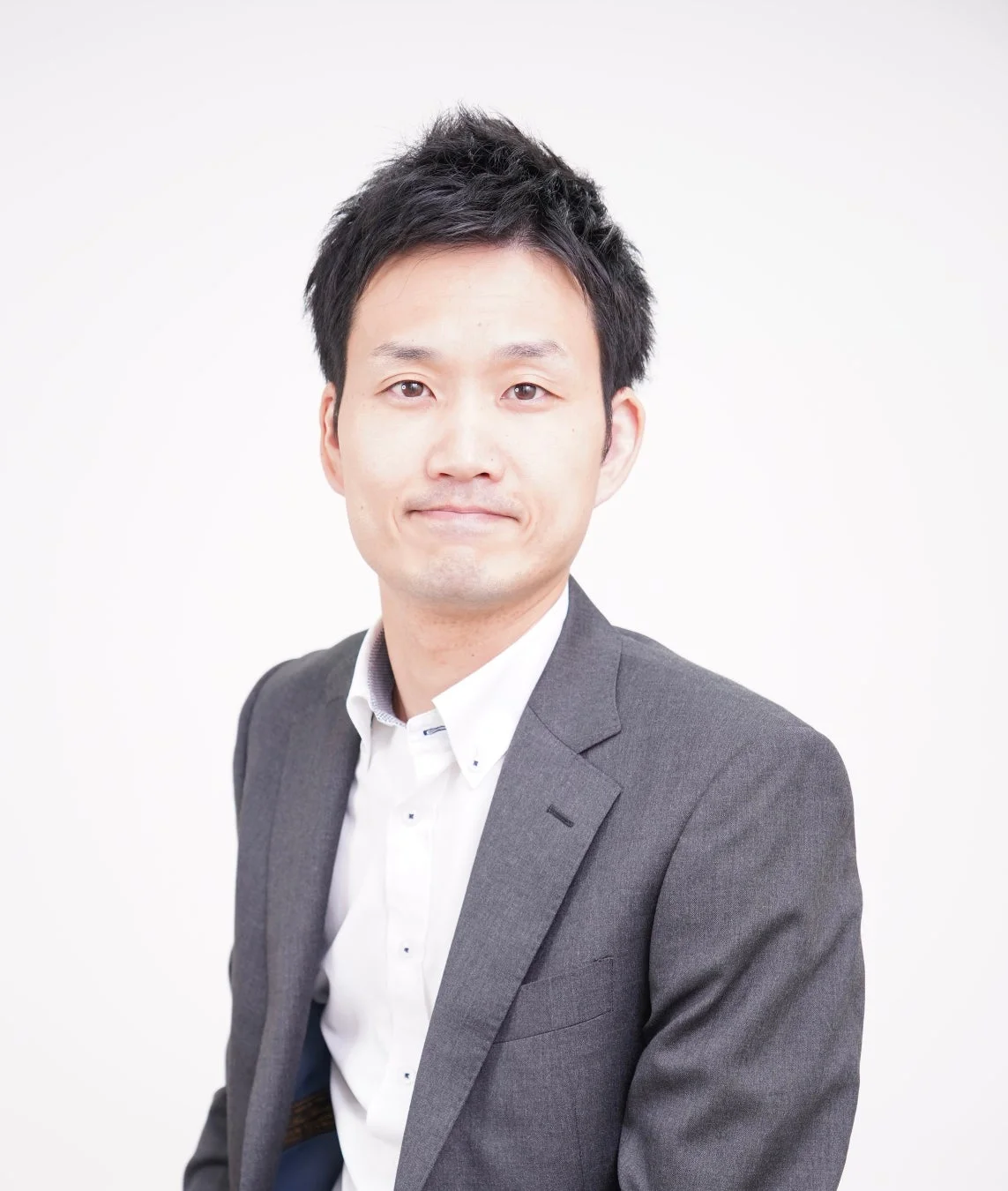 白い背景の前で、グレーのスーツと白いシャツを着用したアジア系の若い男性が、カメラに向かって穏やかな笑顔を見せているポートレート写真