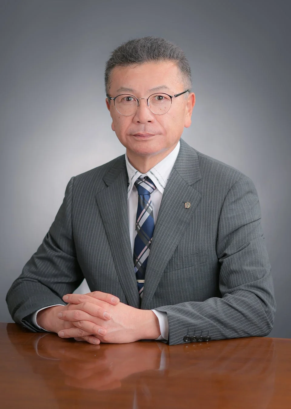 グレーのストライプスーツを着用し、眼鏡をかけたアジア人男性が木製のテーブルに座り、手を組んでカメラを見ている肖像写真