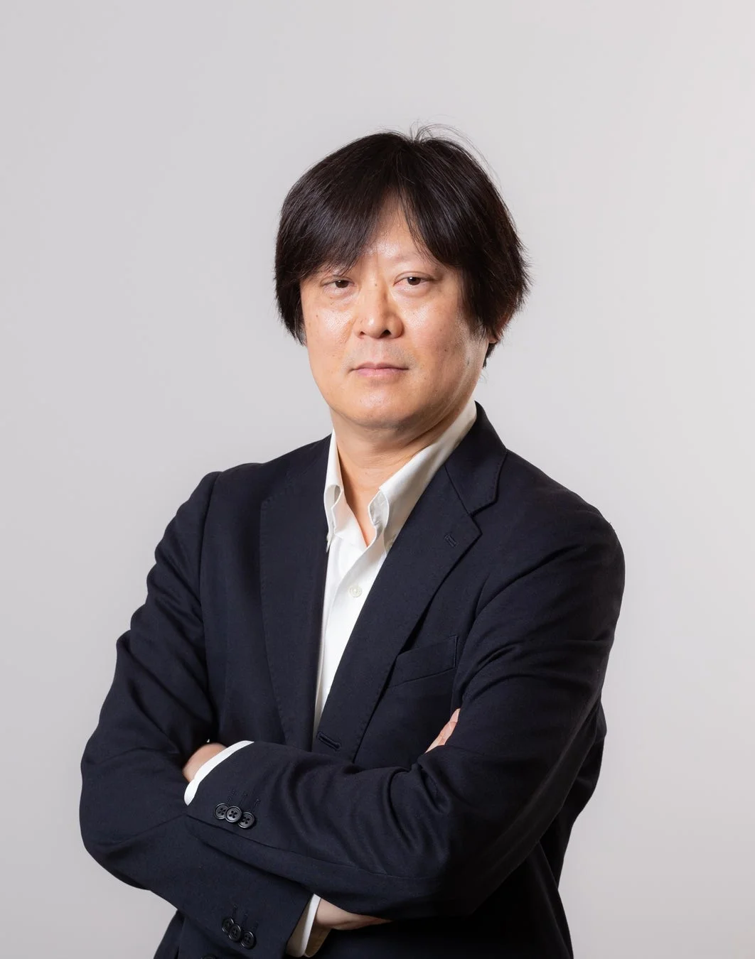 白い背景の前で、濃い色のスーツと白いシャツを着用した中年のアジア人男性が腕を組み、カメラをまっすぐ見ているビジネスポートレート