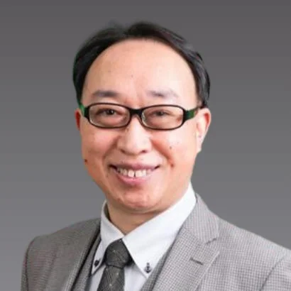 グレーの背景を背に、眼鏡をかけたアジア系の中年男性がスーツとネクタイを着用し、笑顔でカメラを見ている上半身のポートレート画像です。